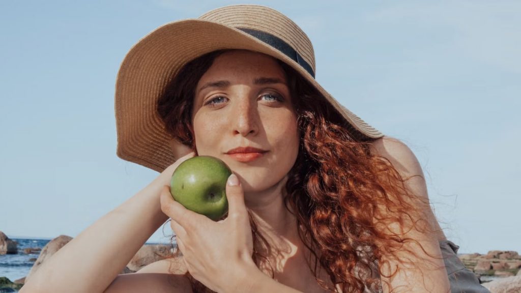 A relaxed woman with a hat holds a green apple, enjoying a sunny beach day.